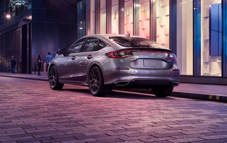 Rear driver-side view of the 2023 Honda Civic Sport Touring Hatchback in Lunar Silver Metallic, parked outside a store.