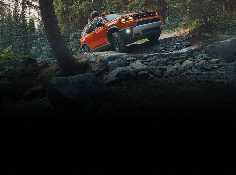 2026 Honda Passport TrailSport Elite in Sunset Orange front off-roading on a rugged rocky forest trail (Mid-Size SUV).